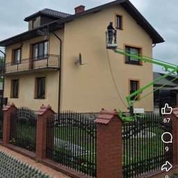 PSWG Czyszczenie Specjalistyczne , remonty i wykończenia - Osoba na zielonym podnośniku koszowym czyści jasnobeżową elewację dwupiętrowego domu z ciemnym dachem i ozdobnym balkonem, otoczonego metalowym płotem.