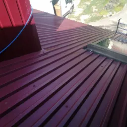 Czerwony dach z blachy trapezowej z zamontowanym oknem dachowym, drabina oparta o okno, widoczna lina zabezpieczająca i fragment samochodu dostawczego w tle.