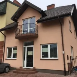 Dwupiętrowy budynek z balkonem i nową, jasnoróżową elewacją, widoczny z poziomu ulicy, z szarą nawierzchnią przed wejściem i ciemnoszarym samochodem zaparkowanym w pobliżu.