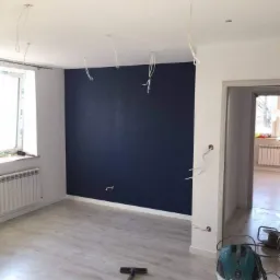 Pok&oacute;j po malowaniu jednej ze ścian na ciemny granat, widoczne wystające kable ze ściany i sufitu, odkurzacz budowlany na podłodze.