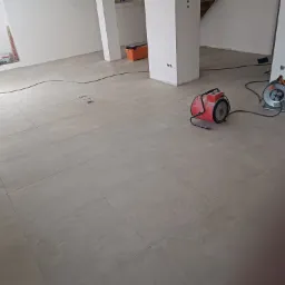 Ułożona jasnoszara podłoga z płytek w surowym wnętrzu, widoczne narzędzia: nagrzewnica, przedłużacz, poziomica i skrzynka narzędziowa.