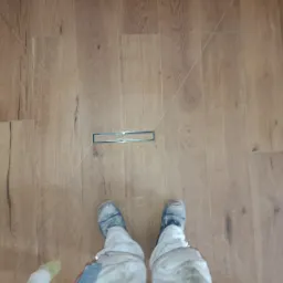 Ułożona podłoga z paneli imitujących drewno, z odpływem liniowym w kabinie prysznicowej. Widoczne stopy osoby w roboczym ubraniu, wykonującej prace wykończeniowe.