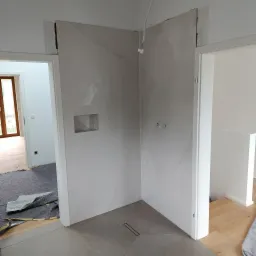 Wykończona kabina prysznicowa z szarymi płytkami imitującymi marmur, wnęką i odpływem liniowym. Widoczne przygotowane instalacje. Dwa wejścia do pomieszczenia.