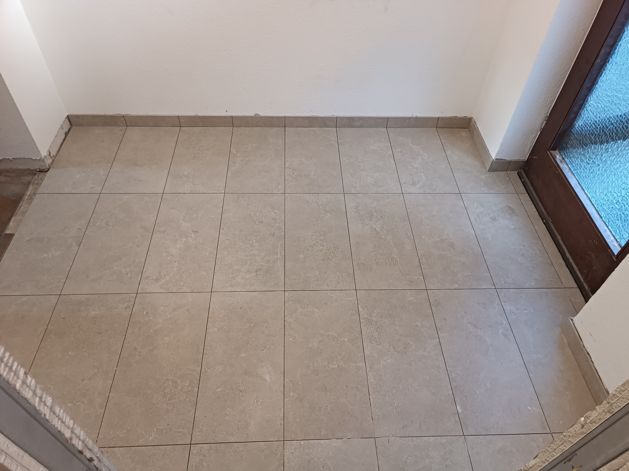 Wyłożona beżowymi płytkami podłoga w korytarzu, z widocznymi fragmentami ściany i drzwi z szybą ornamentową.