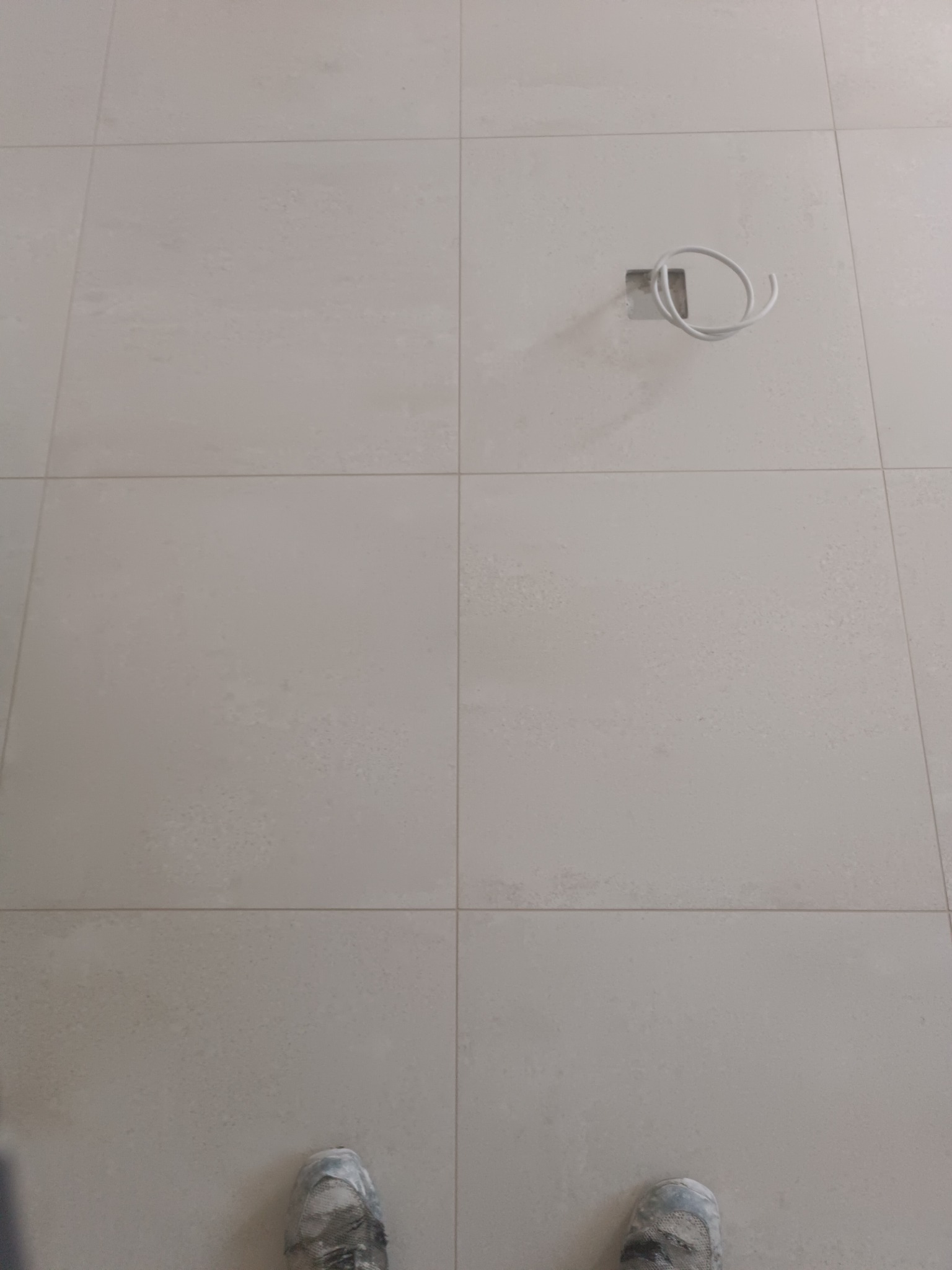 Jasne, matowe płytki podłogowe z widocznym przewodem elektrycznym wystającym ze ściany i fragmentem butów w dolnej części kadru.