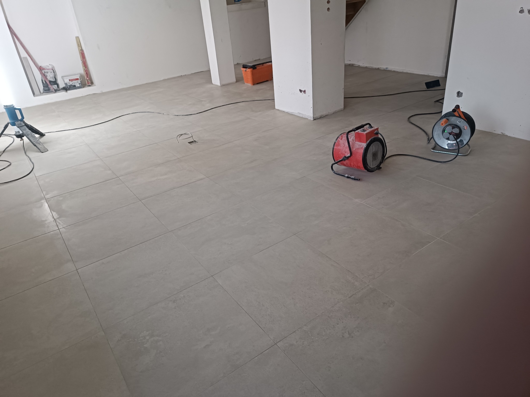 Ułożona jasnoszara podłoga z płytek w surowym wnętrzu, widoczne narzędzia: nagrzewnica, przedłużacz, poziomica i skrzynka narzędziowa.