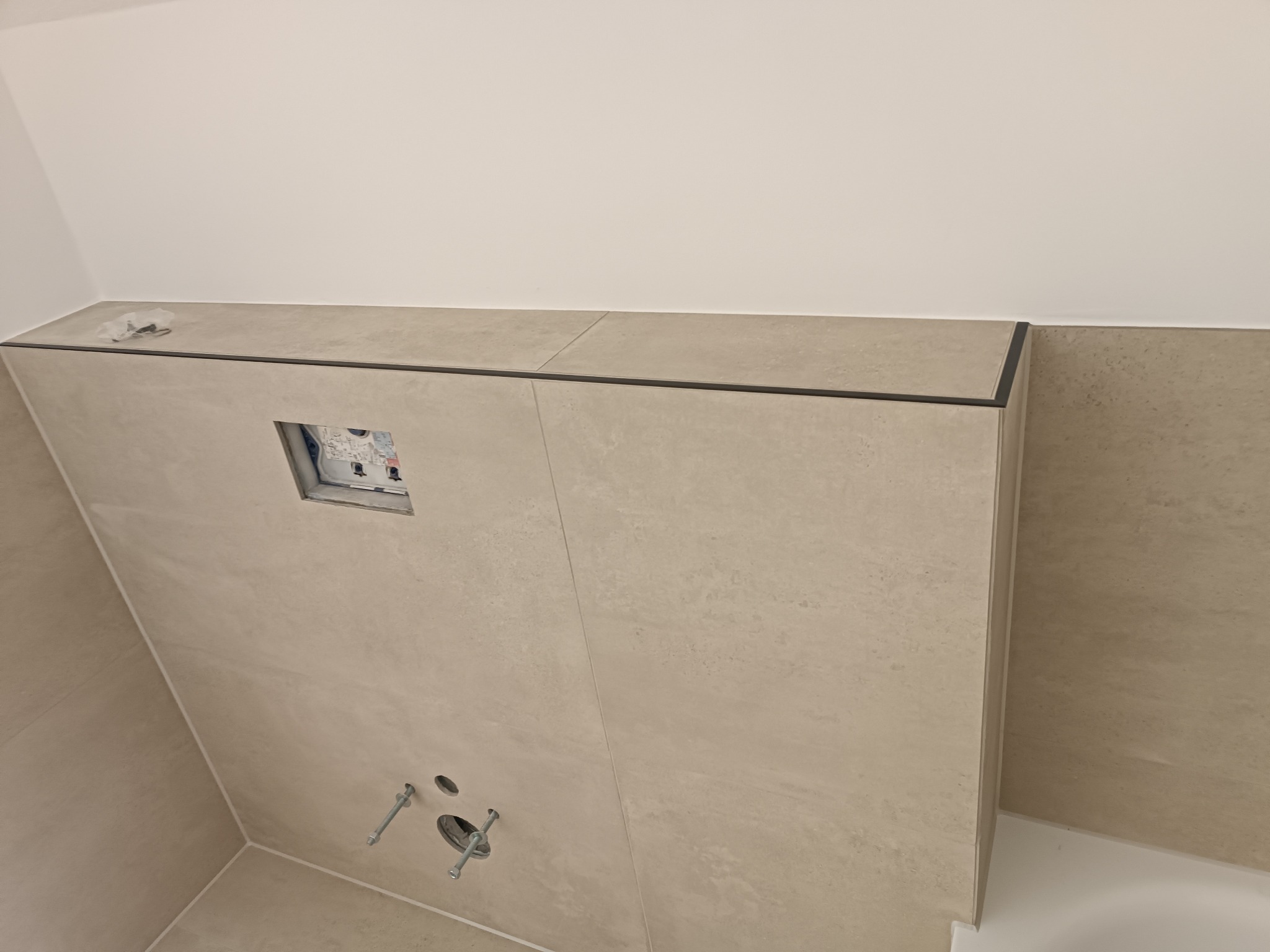 Ściana łazienki wyłożona beżowymi płytkami ceramicznymi z widocznymi elementami instalacji podtynkowych i czarnym profilem wykończeniowym, w trakcie prac remontowych.