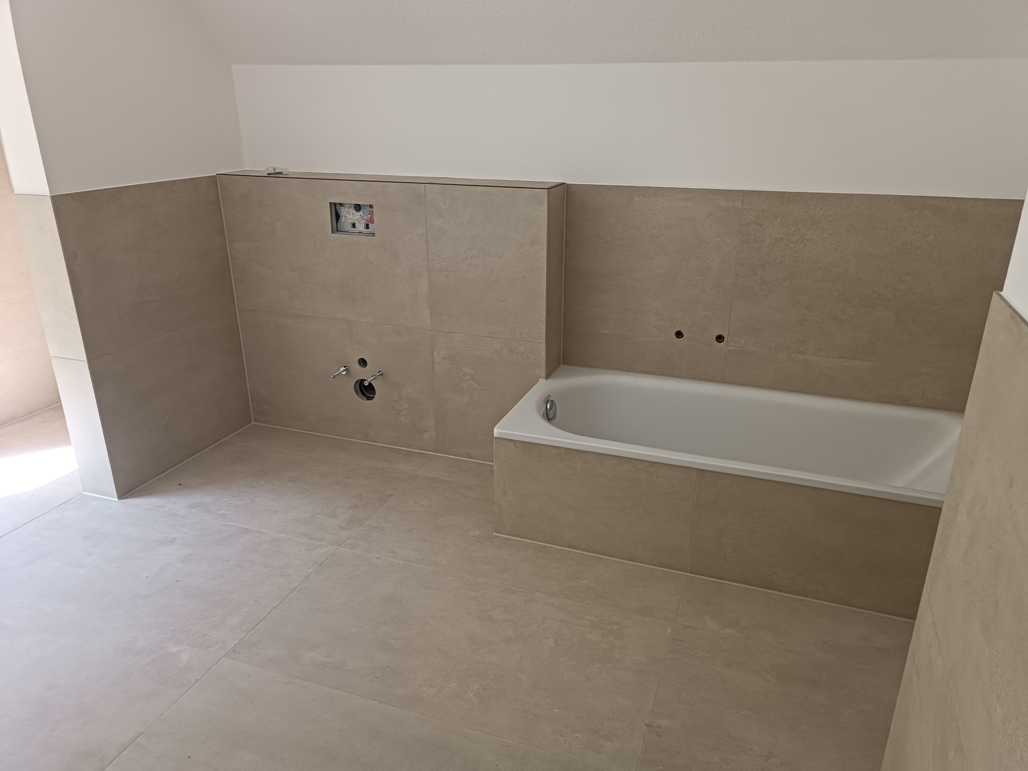 Niedokończona łazienka z beżowymi płytkami podłogowymi i ściennymi, przygotowane otwory na baterie i syfon, oraz zamontowana wanna akrylowa.