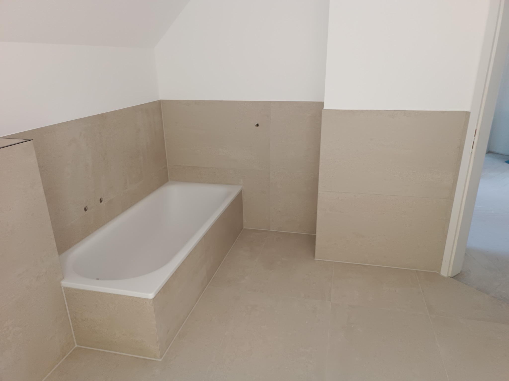 Wykończona łazienka z beżowymi płytkami na ścianach i podłodze, biała wanna zabudowana płytkami, widoczne otwory na baterię w ścianie.