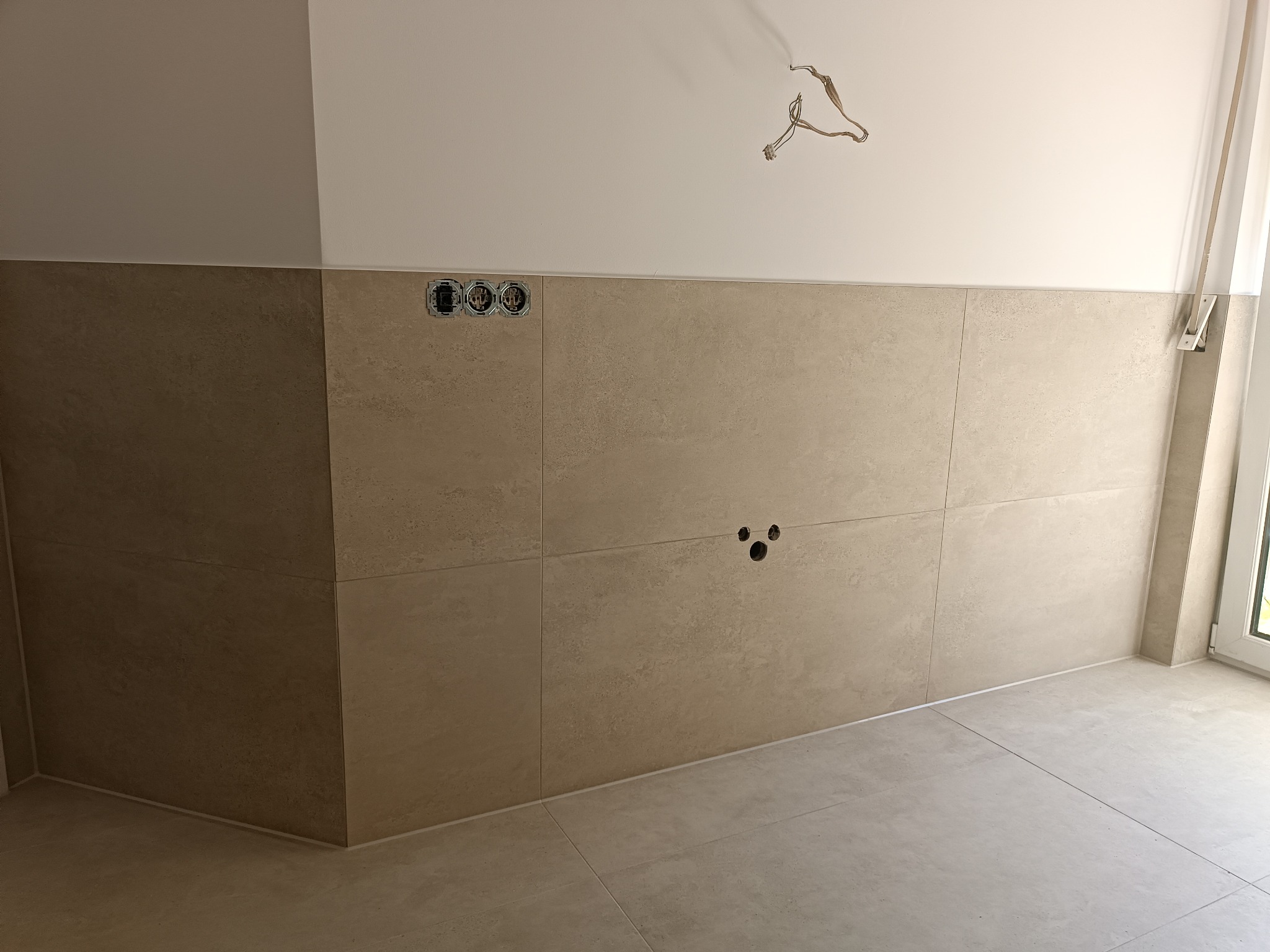 Ściana wyłożona beżowymi płytkami w łazience, widoczne puszki elektryczne i otwory na rury, surowe przewody zwisające z sufitu.