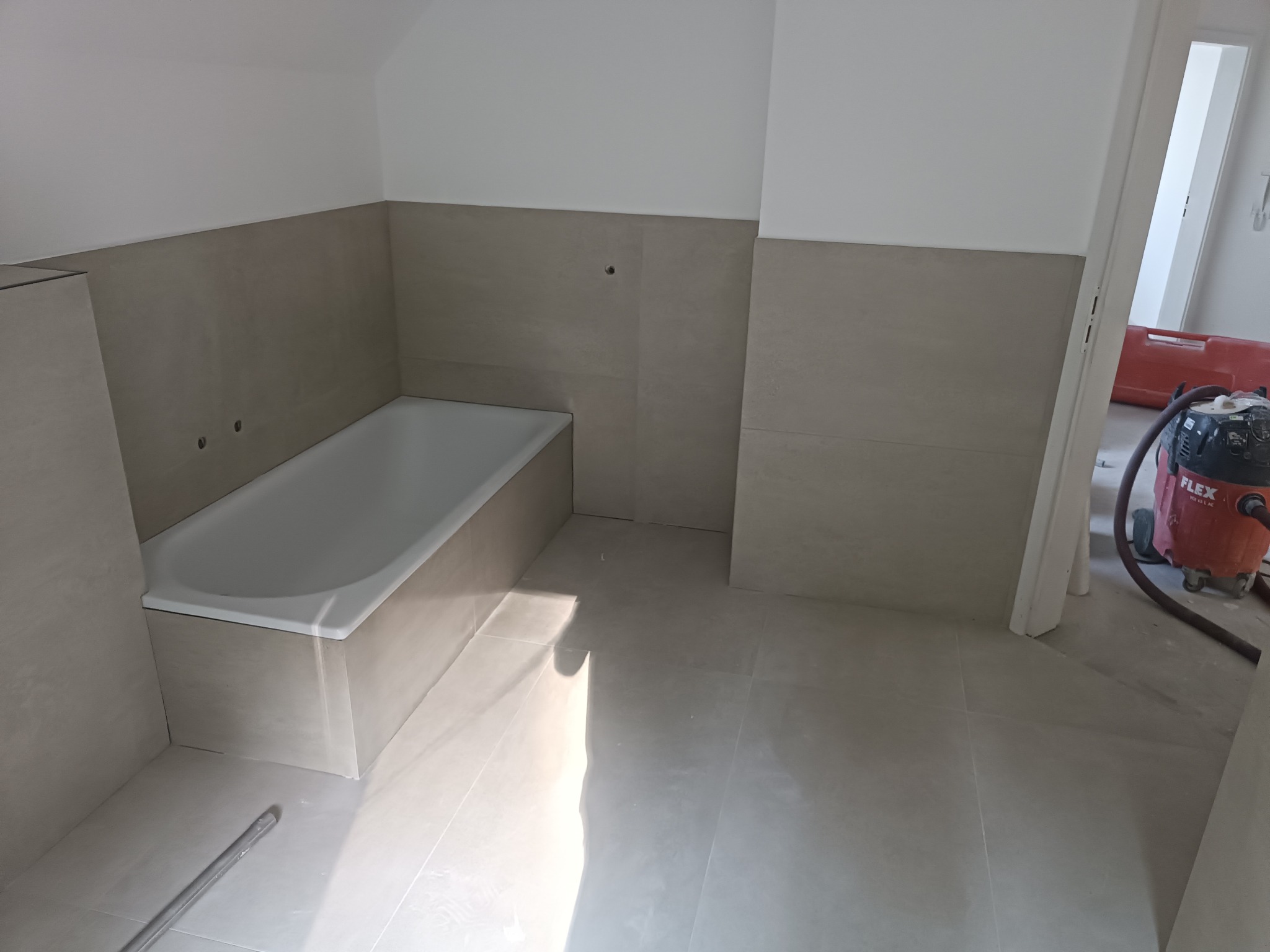 Wanna obudowana płytkami w trakcie prac wykończeniowych; widoczne niedokończone fugi i odkurzacz przemysłowy w tle.