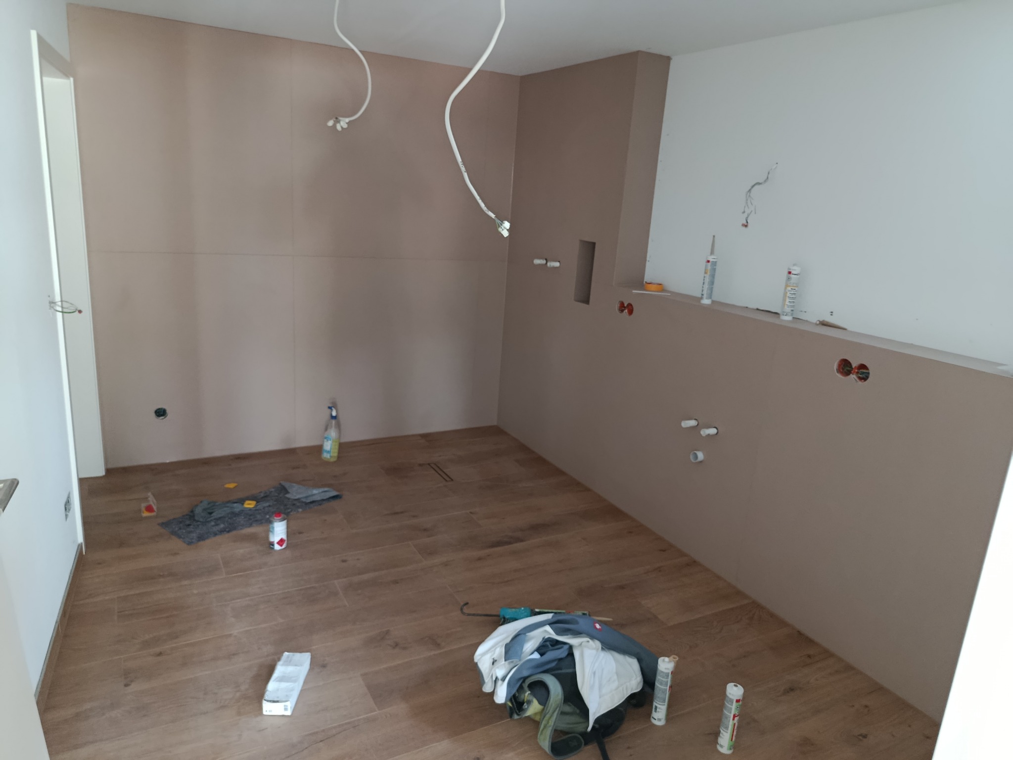 Kuchnia w trakcie remontu: szpachlowane ściany w beżowym odcieniu, drewnopodobne płytki na podłodze, widoczne instalacje i narzędzia, przygotowanie do dalszych prac wykończeniowych.