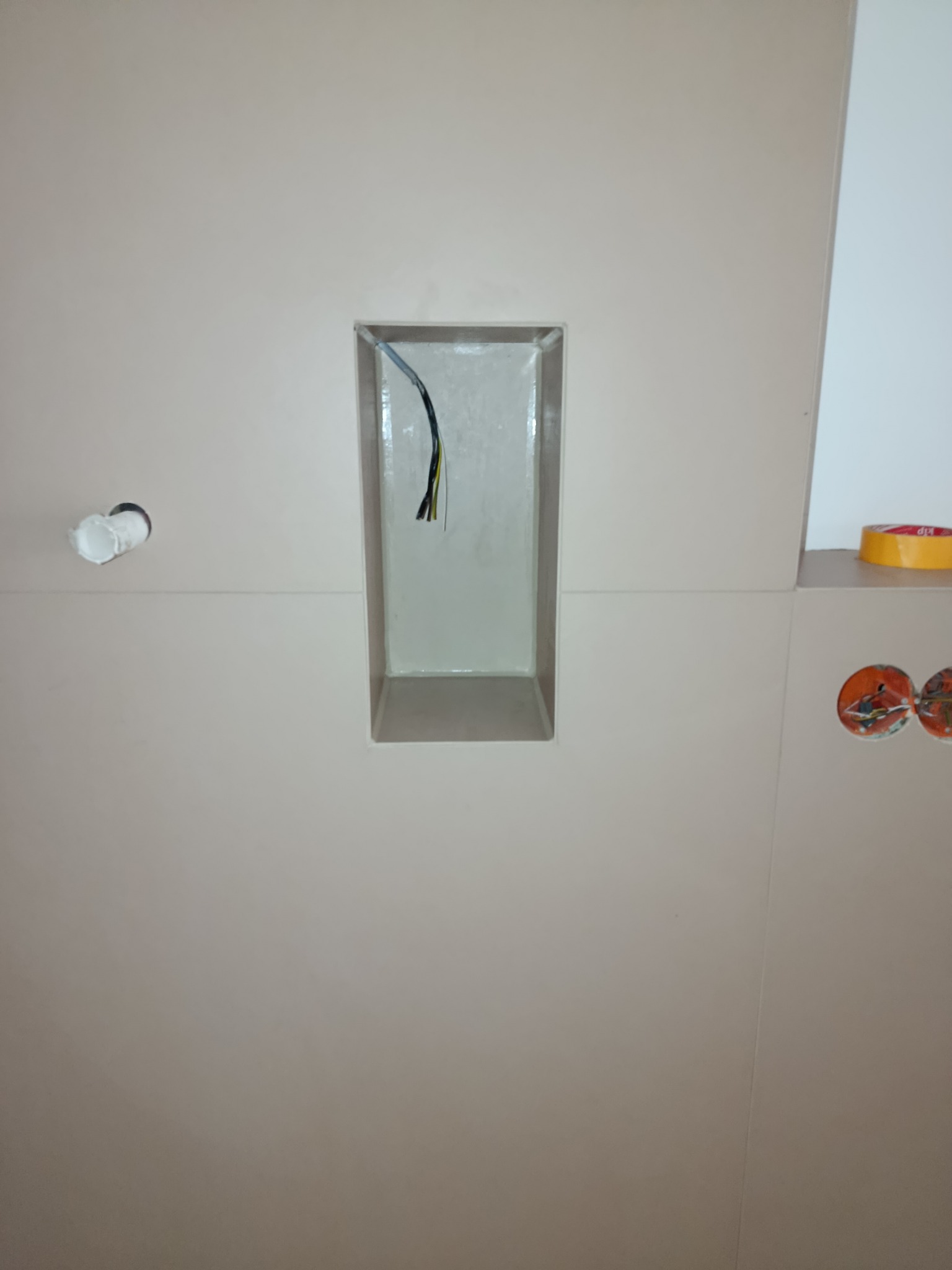 Wykończona ściana z beżowych płytek, wnęka z kablami i instalacja elektryczna z pomarańczowymi puszkami. Widoczna taśma malarska.