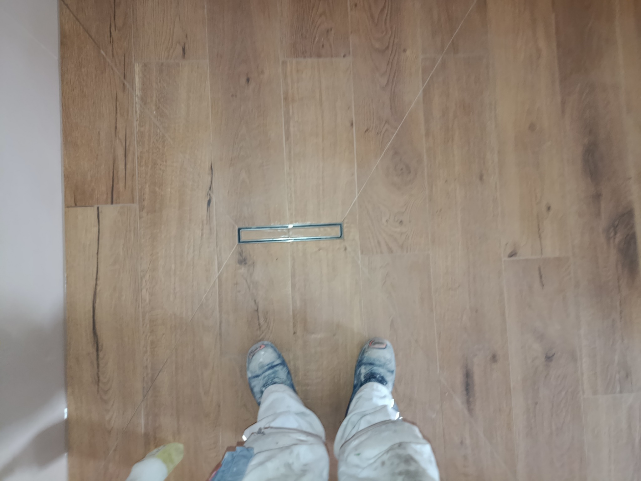 Ułożona podłoga z paneli imitujących drewno, z odpływem liniowym w kabinie prysznicowej. Widoczne stopy osoby w roboczym ubraniu, wykonującej prace wykończeniowe.