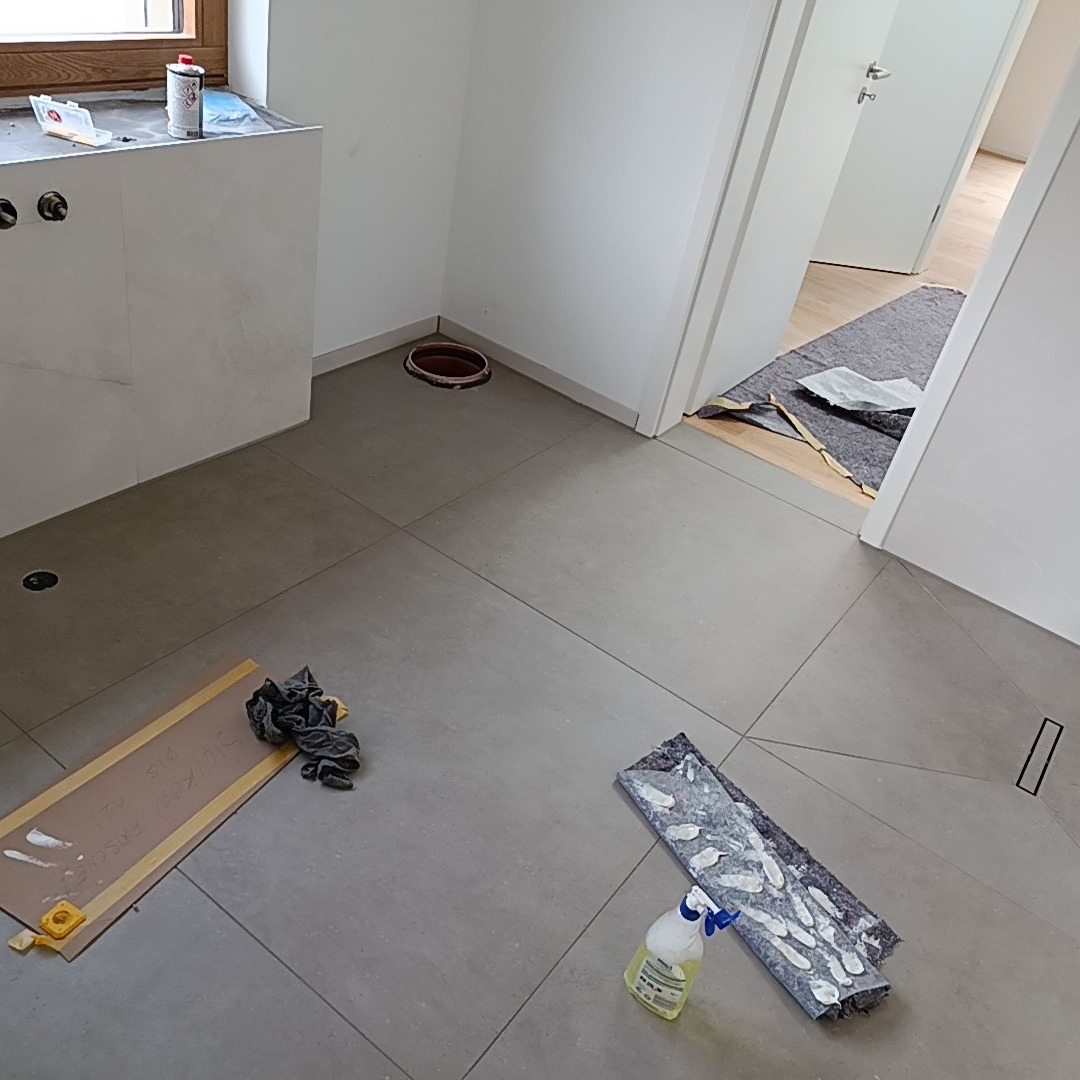 Szare płytki podłogowe w trakcie układania, widoczne narzędzia glazurnicze i fragment umywalki. Otwór kanalizacyjny i przejście do innego pomieszczenia.