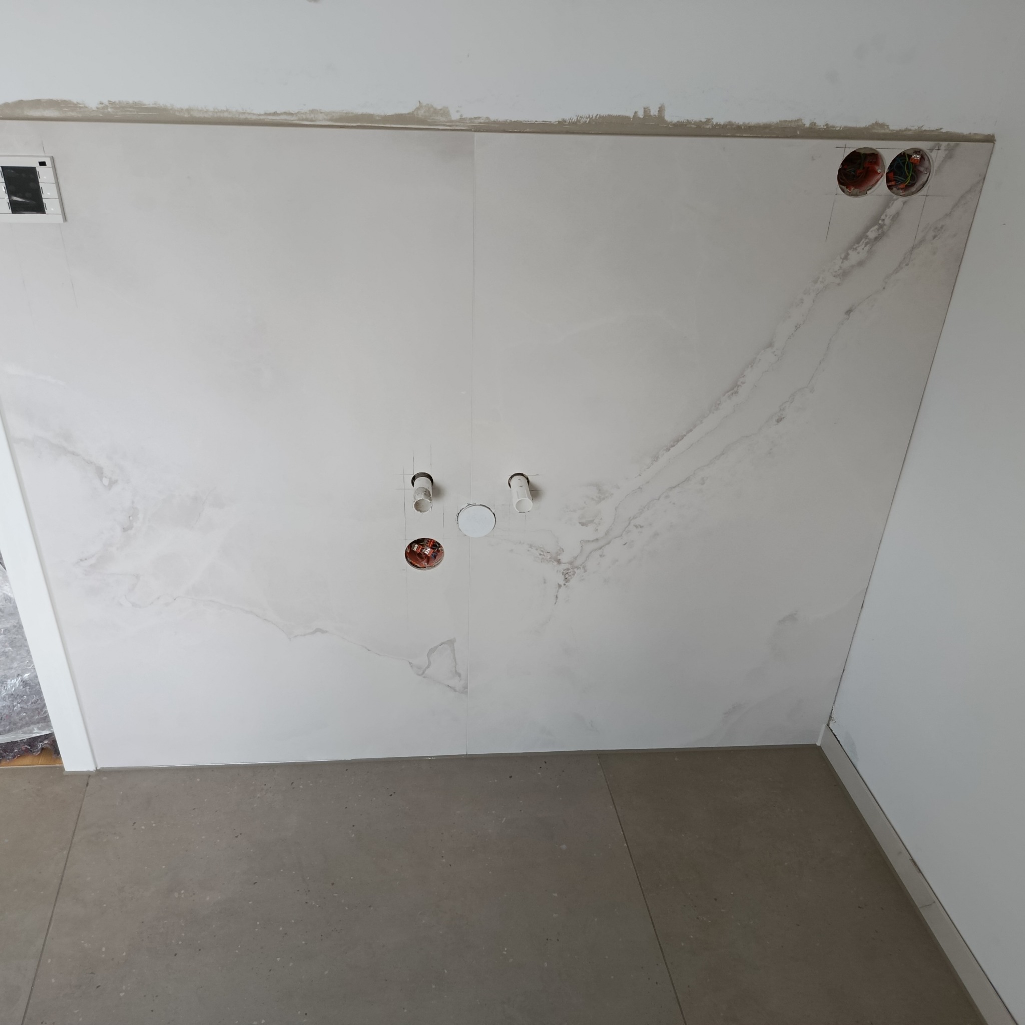 Ściana w łazience wyłożona płytkami imitującymi marmur, z widocznymi otworami na instalacje wodno-kanalizacyjne i elektryczne. Szare płytki podłogowe.