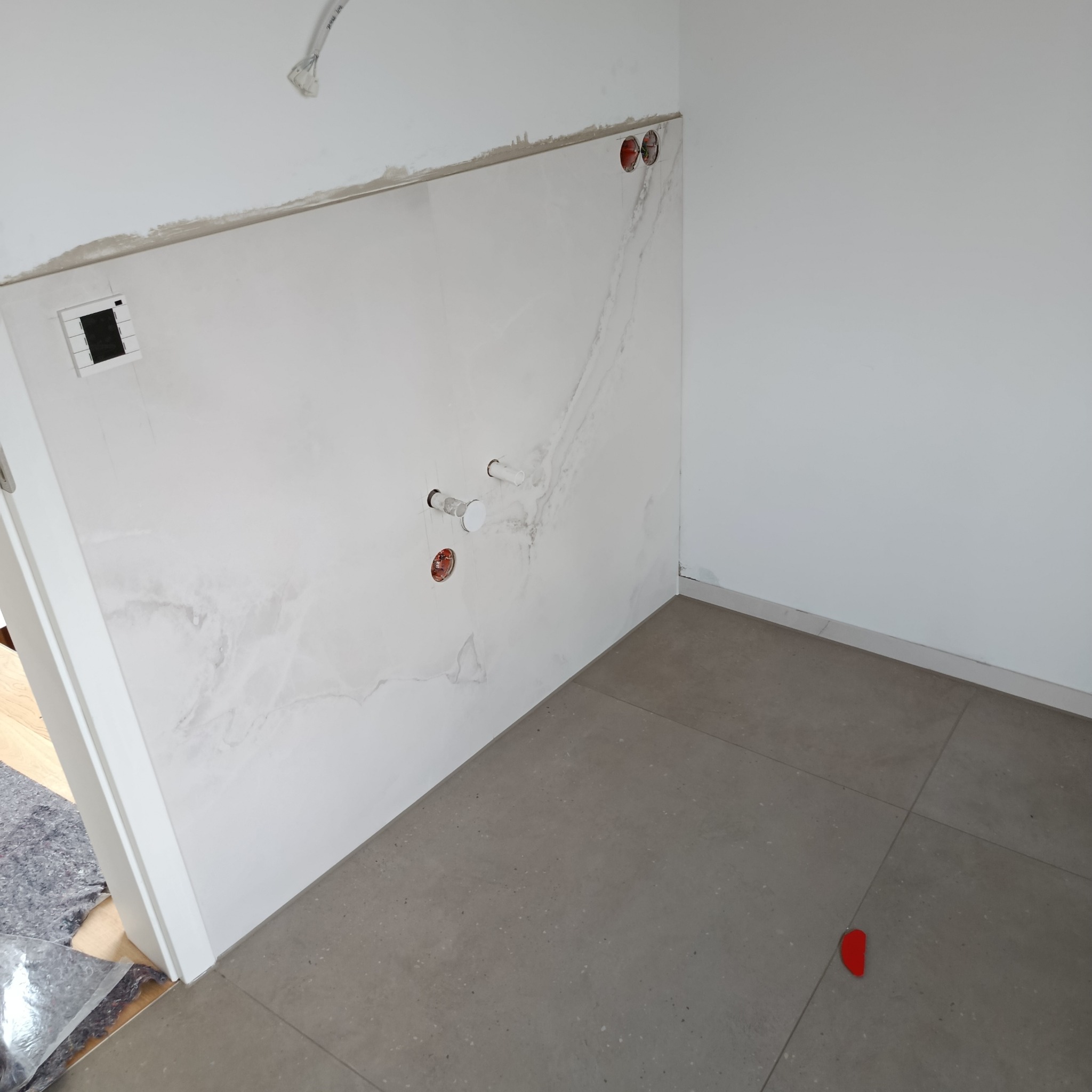 Fragment wykończonej łazienki z marmurową płytą na ścianie, widoczne otwory na instalacje i fragment szarej podłogi z płytek. W tle biała ściana i fragment drzwi.