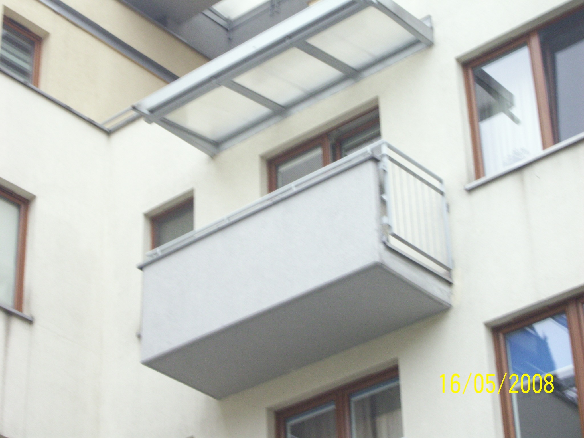 Balkon z szarą zabudową i zadaszeniem z poliwęglanu, widok z dołu na tle elewacji budynku z oknami w brązowych ramach. Na zdjęciu widoczna data 16/05/2008.
