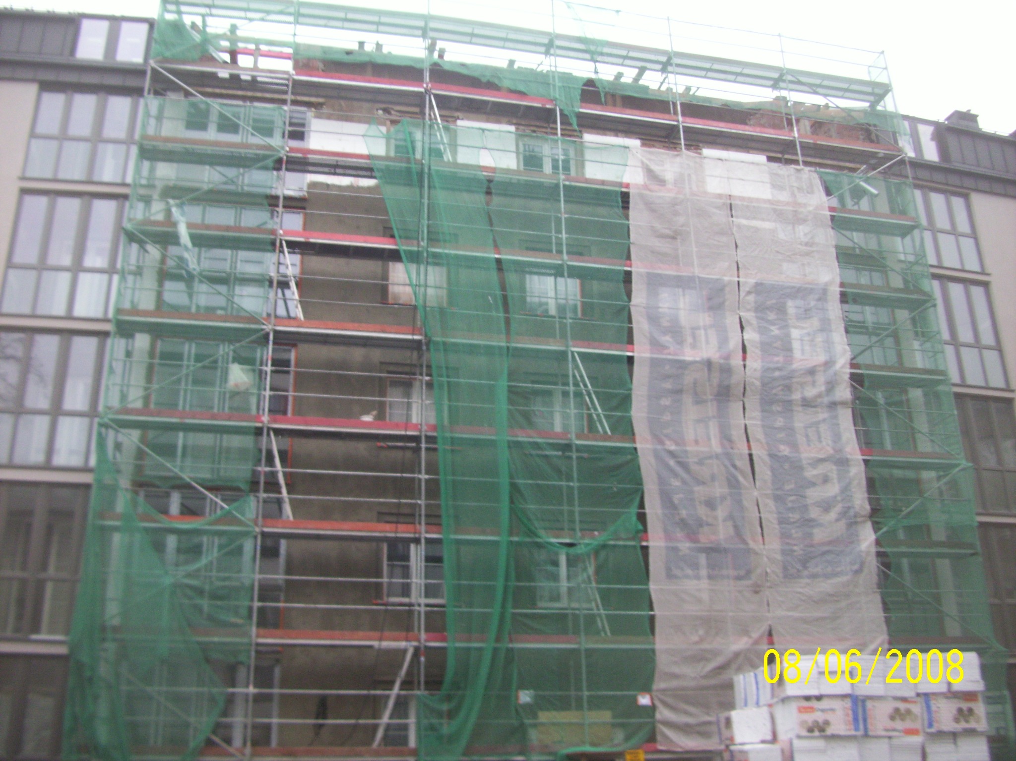 Budynek w trakcie renowacji z rusztowaniami, siatką ochronną i materiałami budowlanymi u podstawy, widoczny fragment elewacji bez tynku.