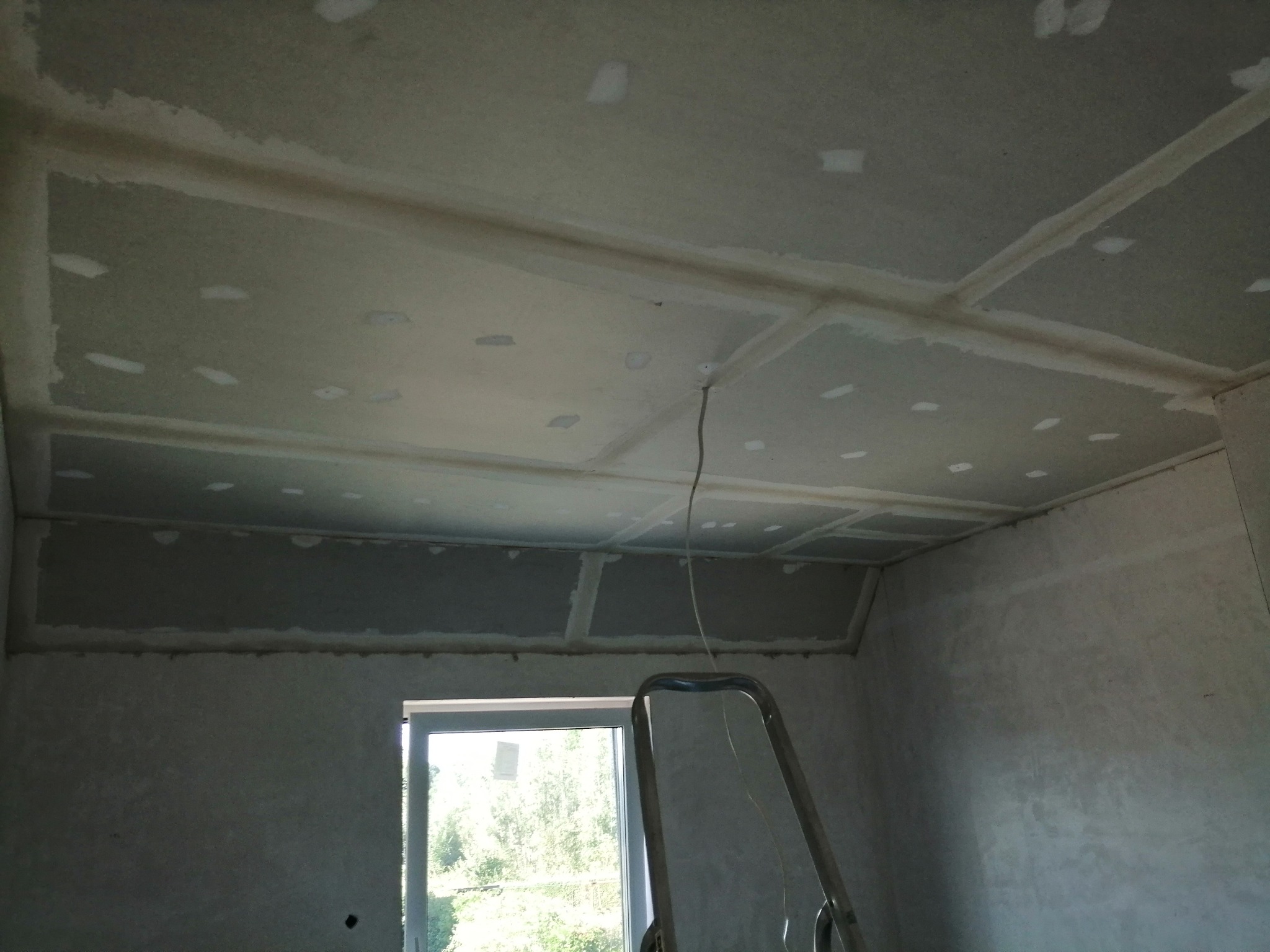 Sufit z wykończonymi łączeniami płyt, widoczne szpachlowane miejsca na wkręty, drabina oparta o ścianę, okno z widokiem na zieleń.