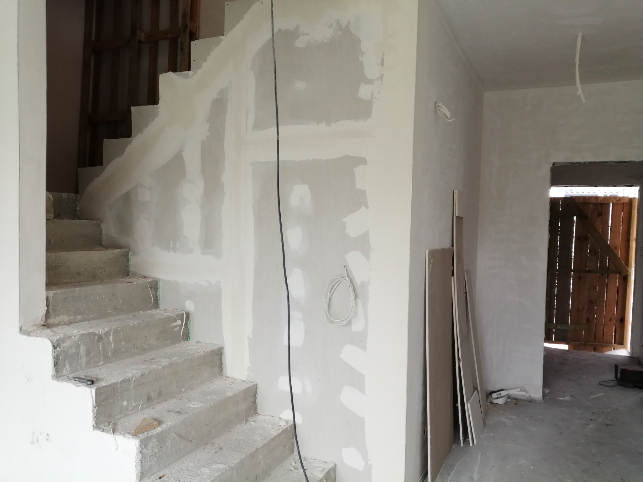 Montaż okładziny na ścianie przy schodach, widoczne surowe betonowe schody, kabel elektryczny zwisający ze ściany, przygotowane płyty do montażu, częściowo wykończone powierzchnie.