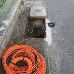 OPTECH_Teletechnika, Elektryka, Klimatyzacja - Pomarańczowy kabel telekomunikacyjny zwinięty w krąg leży obok otwartej studzienki kanalizacyjnej, z której wystają inne kable.