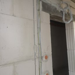 OPTECH_Teletechnika, Elektryka, Klimatyzacja - Świeżo poprowadzone kable elektryczne w białej ścianie z betonu komórkowego, widoczne puszki instalacyjne i zwój kabli pod sufitem przy przejściu.