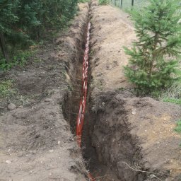 OPTECH_Teletechnika, Elektryka, Klimatyzacja - Wykop w ziemi z pomarańczowymi rurami telekomunikacyjnymi ułożonymi wzdłuż ogrodzenia z siatki.