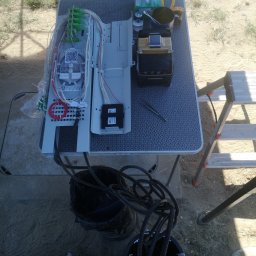 OPTECH_Teletechnika, Elektryka, Klimatyzacja - Zestaw do spawania światłowodów na składanym stole z wzorem geometrycznym, widoczne złącza, spawarka, akcesoria i drabina w tle na zewnątrz.