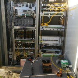 OPTECH_Teletechnika, Elektryka, Klimatyzacja - Otwarta szafa telekomunikacyjna z rozbudowanym okablowaniem światłowodowym i urządzeniami sieciowymi, narzędzia i akcesoria na rozkładanym stoliku przed szafą, widoczne złącza, patch panele...