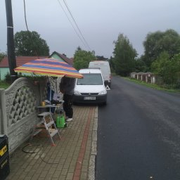 OPTECH_Teletechnika, Elektryka, Klimatyzacja - Pracownik w trakcie pracy na chodniku, pod kolorowym parasolem, obok zaparkowanego białego busa z trójkątem ostrzegawczym, widoczne narzędzia i sprzęt.