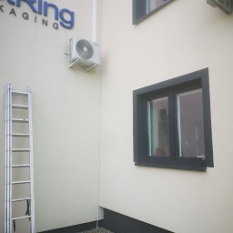 OPTECH_Teletechnika, Elektryka, Klimatyzacja - Zewnętrzny montaż klimatyzacji MDV na elewacji budynku firmy z widoczną drabiną aluminiową i oknami z ciemnymi ramami.