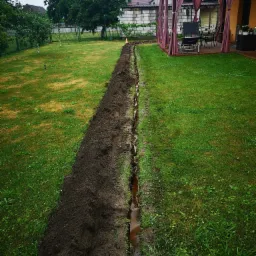 Wykopany r&oacute;w w trawniku, prawdopodobnie pod instalację kablową lub drenażową, z widoczną folią ochronną wewnątrz, prowadzący w kierunku domu z pergolą.