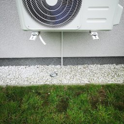 OPTECH_Teletechnika, Elektryka, Klimatyzacja - Biała jednostka zewnętrzna klimatyzacji zamontowana na elewacji budynku, widoczne białe kamienie dekoracyjne i trawa.