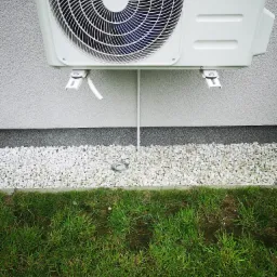 Biała jednostka zewnętrzna klimatyzacji zamontowana na elewacji budynku, widoczne białe kamienie dekoracyjne i trawa.