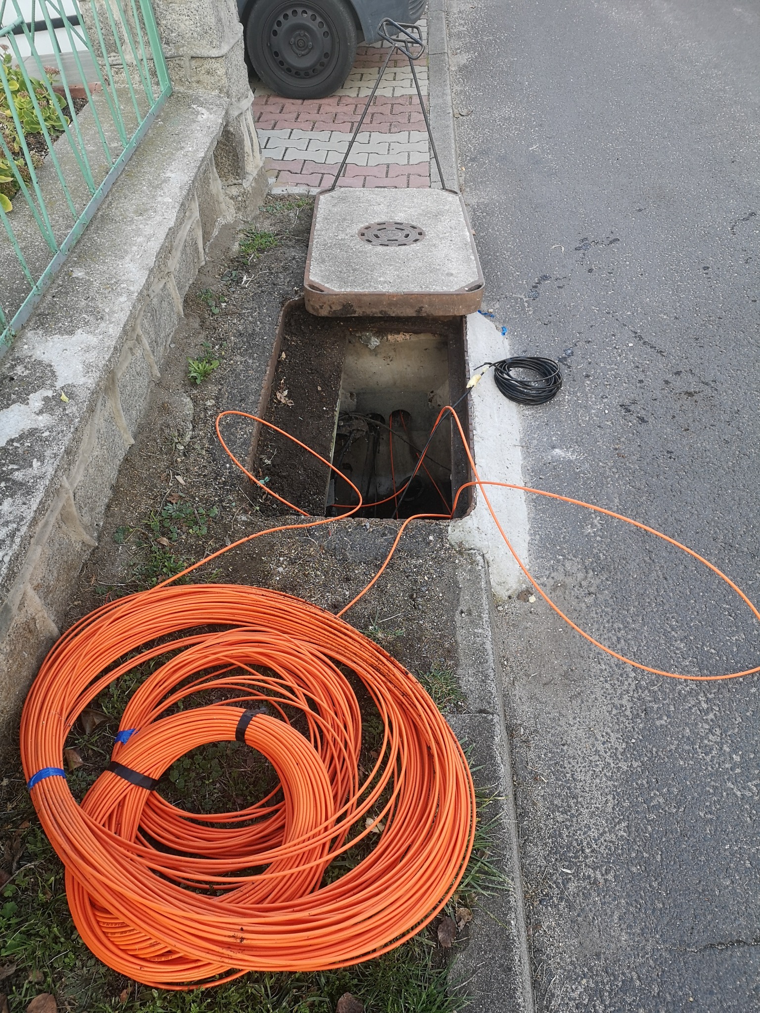 Pomarańczowy kabel telekomunikacyjny zwinięty w krąg leży obok otwartej studzienki kanalizacyjnej, z której wystają inne kable.