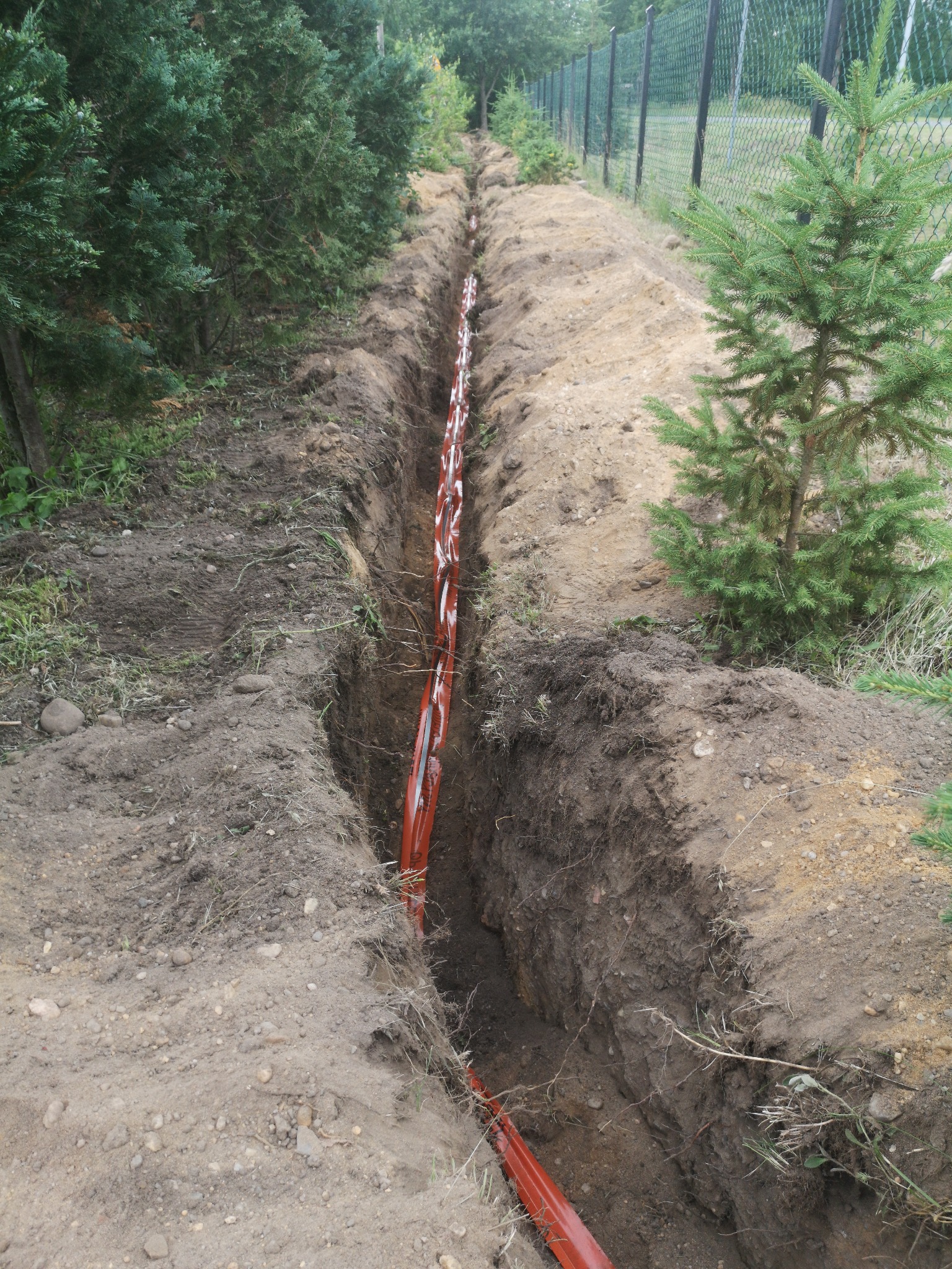 Wykop w ziemi z pomarańczowymi rurami telekomunikacyjnymi ułożonymi wzdłuż ogrodzenia z siatki.