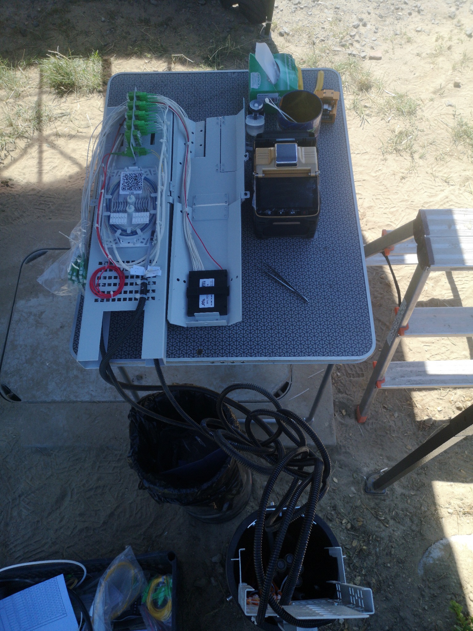 Zestaw do spawania światłowodów na składanym stole z wzorem geometrycznym, widoczne złącza, spawarka, akcesoria i drabina w tle na zewnątrz.