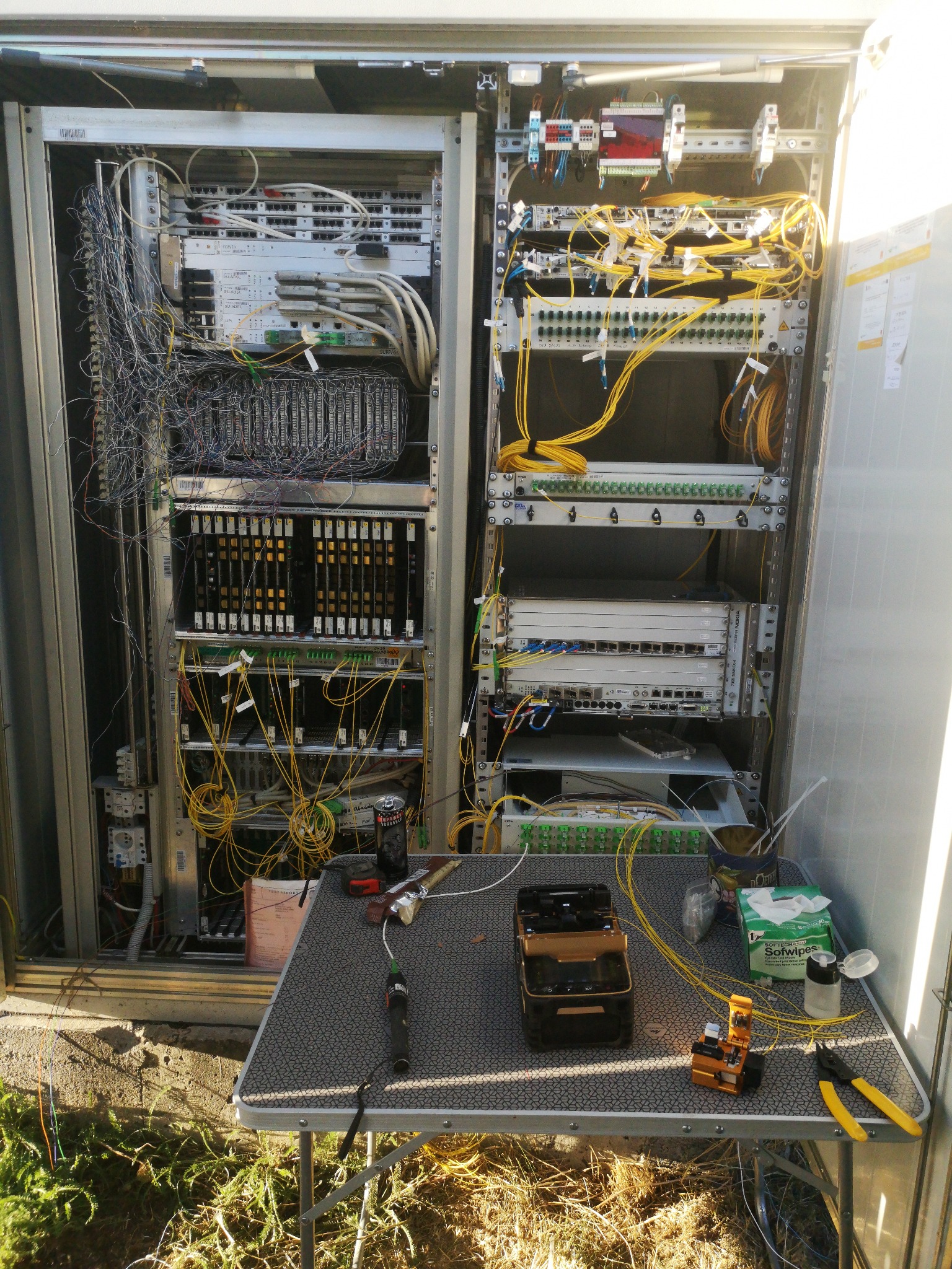 Otwarta szafa telekomunikacyjna z rozbudowanym okablowaniem światłowodowym i urządzeniami sieciowymi, narzędzia i akcesoria na rozkładanym stoliku przed szafą, widoczne złącza, patch panele...