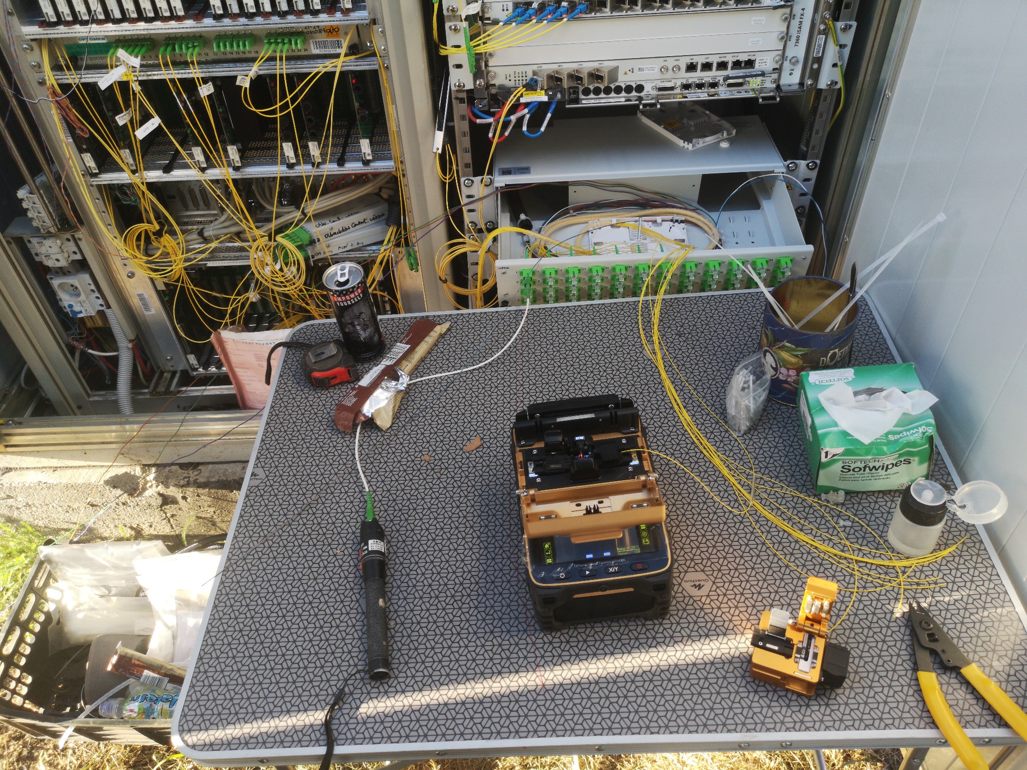 Przenośny stół roboczy z narzędziami do spawania światłowodów, testerem, obcinarkami i zapasem chusteczek higienicznych na tle szafy telekomunikacyjnej z kablem światłowodowym i puszką napoju...