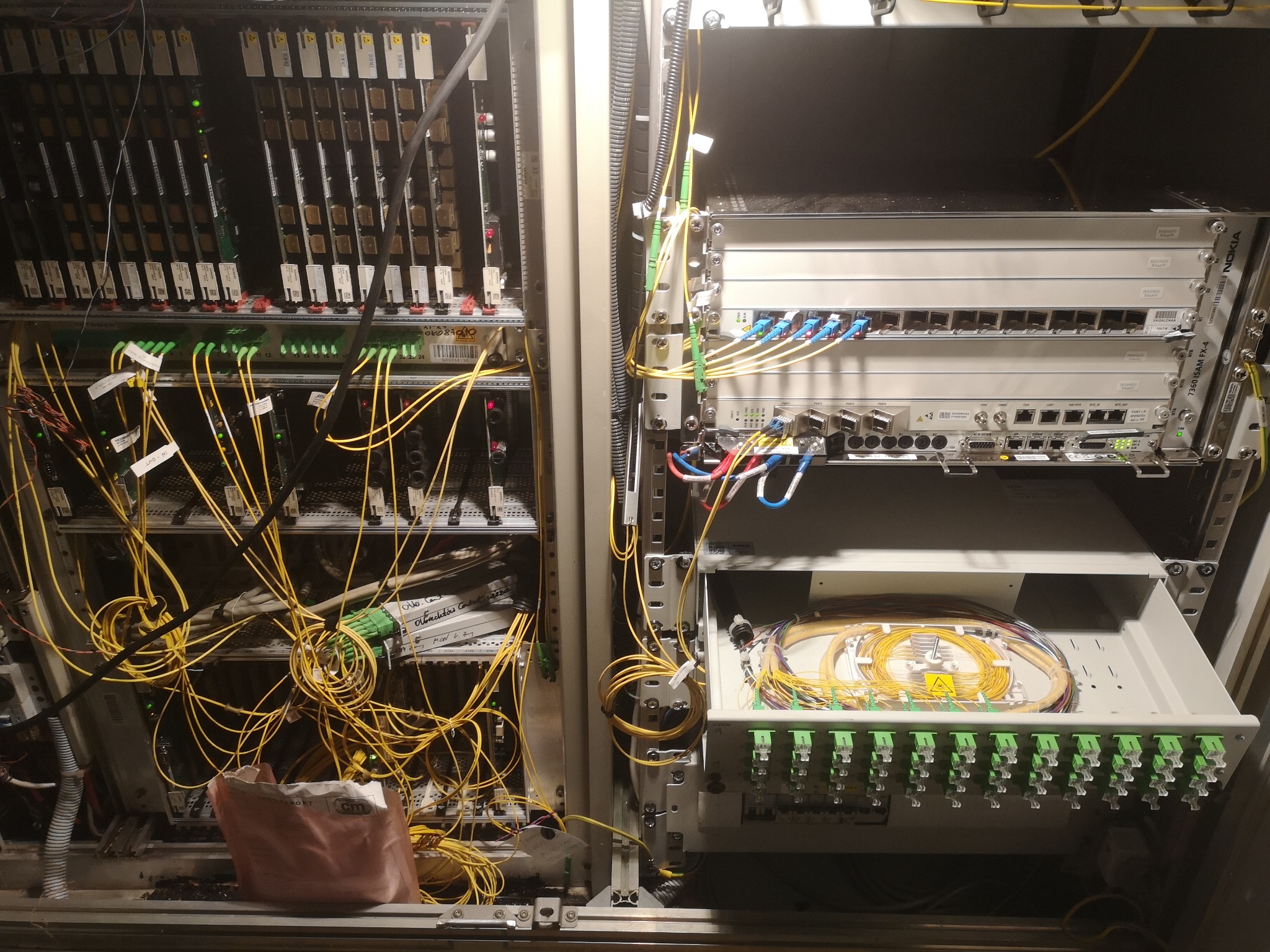 Szafa telekomunikacyjna z widocznymi modułami światłowodowymi, patch panelem i licznymi kablami. W dolnej części szafy widoczny bałagan i torba po materiałach.