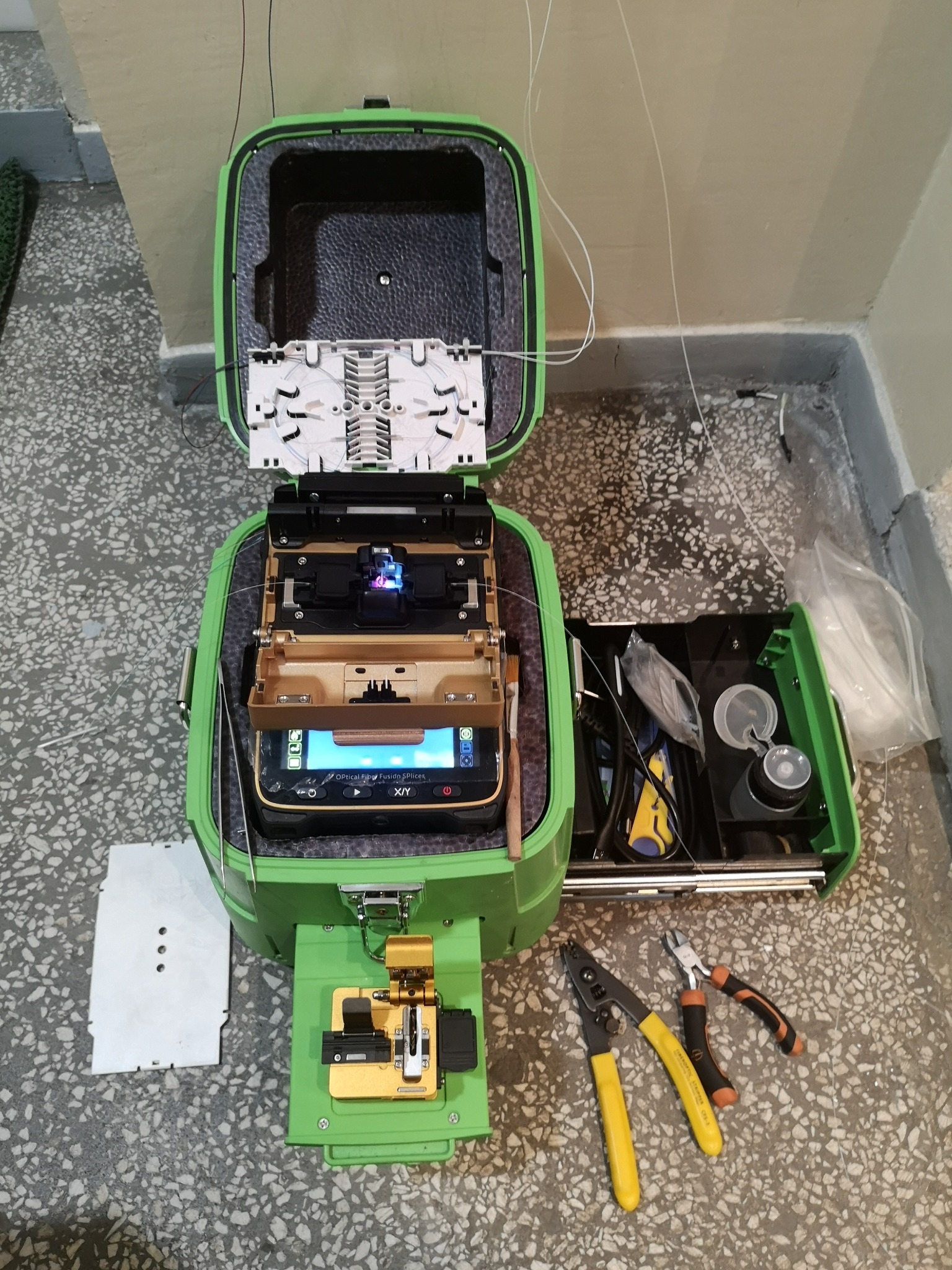 Otwarty, zielony splicer światłowodowy z widocznymi włóknami światłowodowymi, narzędziami i akcesoriami na podłodze z lastryko.