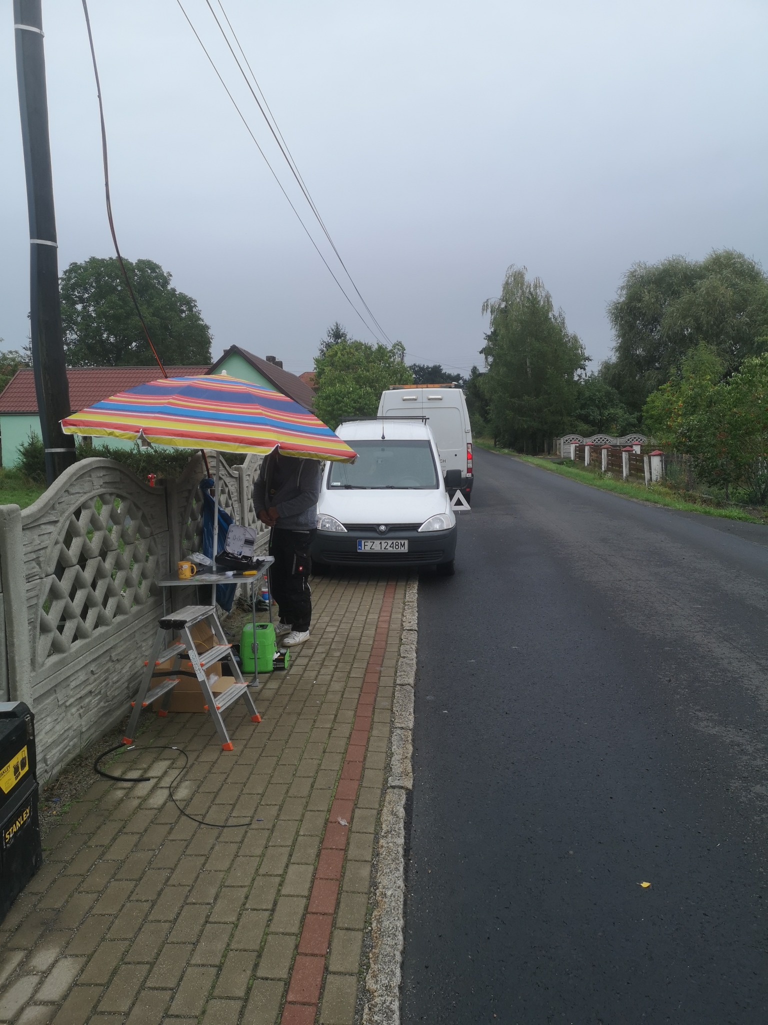Pracownik w trakcie pracy na chodniku, pod kolorowym parasolem, obok zaparkowanego białego busa z trójkątem ostrzegawczym, widoczne narzędzia i sprzęt.