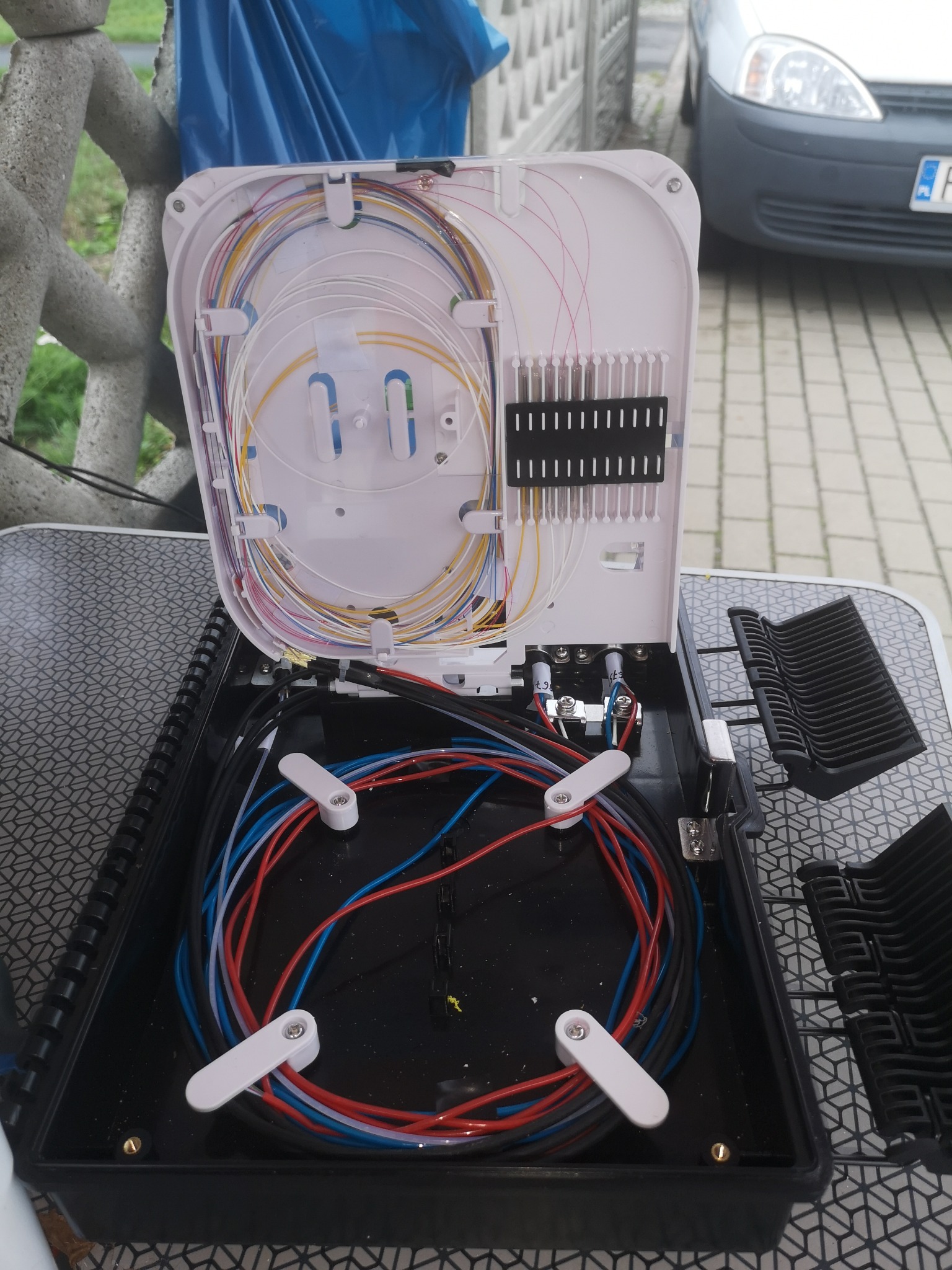 Otwarta skrzynka telekomunikacyjna z rozłożonymi kablami światłowodowymi i przewodami w kolorach: białym, żółtym, czerwonym, niebieskim i czarnym, widoczne złącza i uchwyty, tło: kostka brukowa...