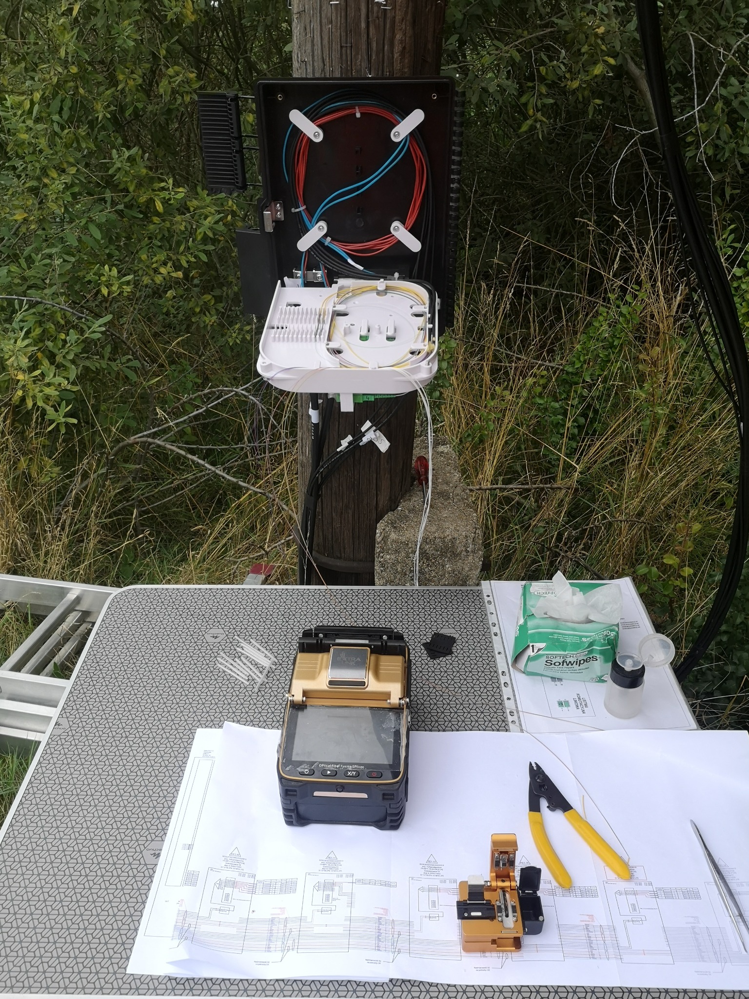 Skrzynka telekomunikacyjna z rozłożonymi kablami światłowodowymi na słupie, z narzędziami do spawania światłowodów i schematem na stole roboczym w tle roślinność.