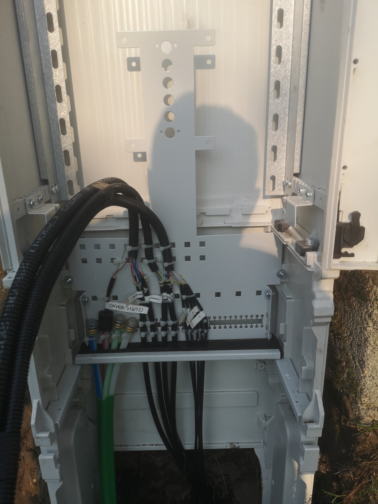 Otwarta szafka telekomunikacyjna z podłączonymi kablami światłowodowymi i ethernetowymi, widoczne oznaczenia OKH087919/12J na etykietach kabli.