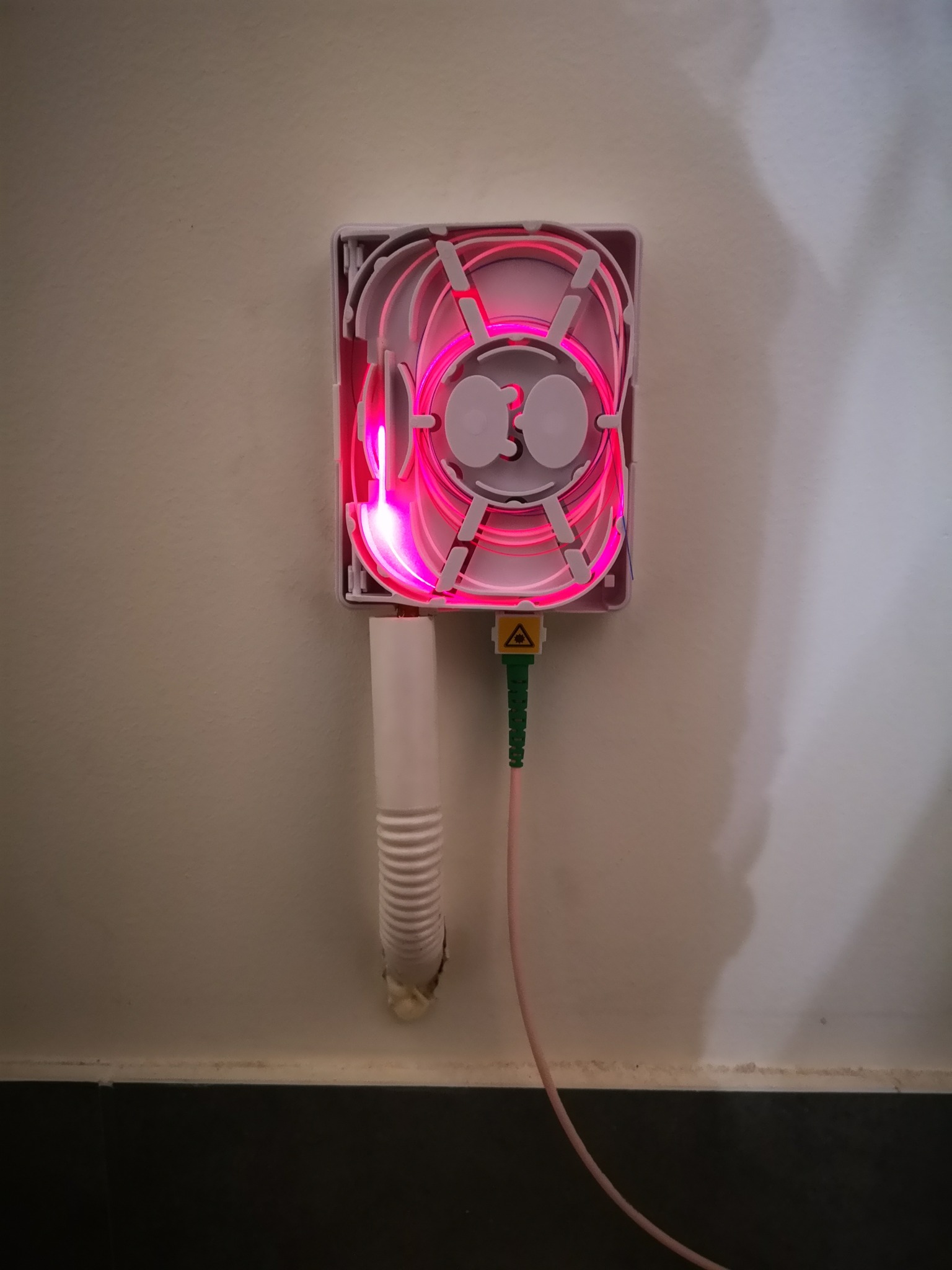 Światłowodowy patch panel z różowym światłem wewnątrz, zamontowany na ścianie, z widocznym kablem światłowodowym i peszlem.