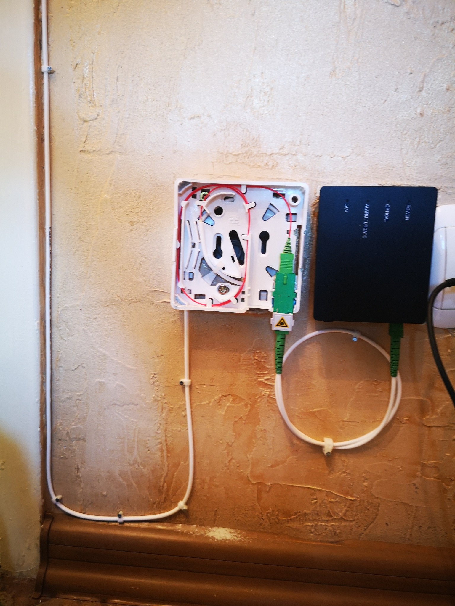 Otwarta puszka telekomunikacyjna z podłączonym światłowodem i czarnym routerem na teksturowanej ścianie, widoczne kable i listwa przypodłogowa.