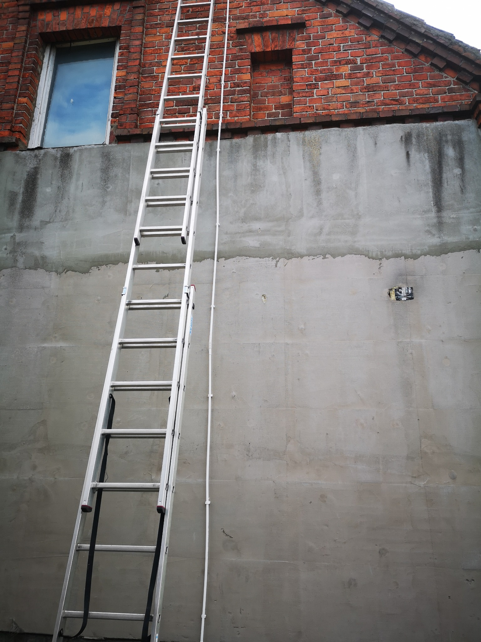 Aluminiowa drabina oparta o szarą, otynkowaną ścianę budynku z czerwonej cegły, widoczny fragment okna z odbiciem nieba.