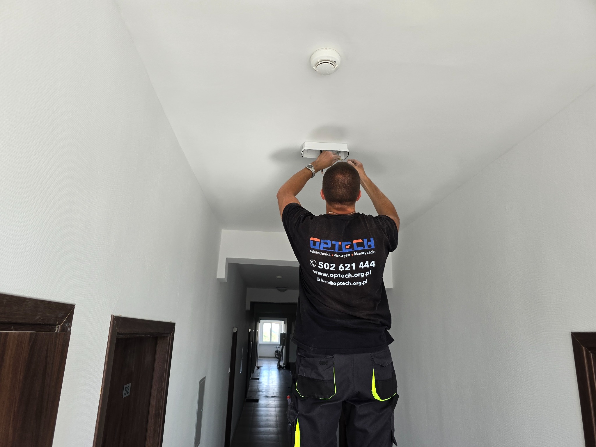Elektryk montuje białą lampę sufitową w korytarzu, widoczny czujnik dymu i logo firmy na koszulce montera.